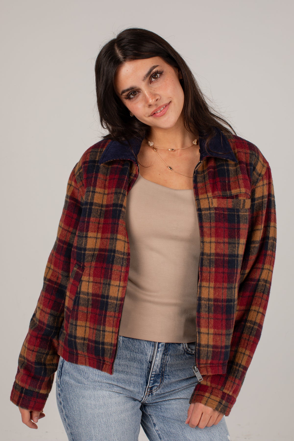 A woman modeling a red and navy plaid flannel shirt jacket over a beige tank top and blue jeans, photographed against a neutral light gray studio background.