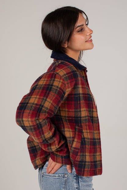 A woman shown in profile wearing a red, navy, and tan plaid flannel shirt jacket over a dark navy collared shirt, paired with light blue jeans, against a plain light gray studio background.