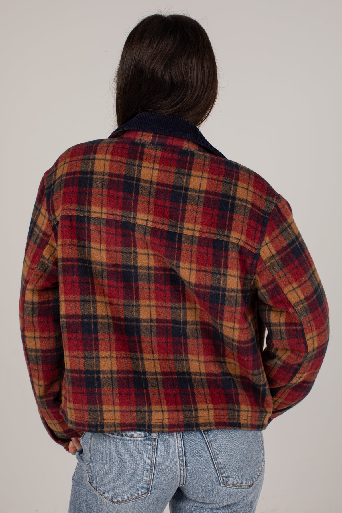 A person viewed from the back wearing a red, navy, and tan plaid flannel-style jacket with a dark navy collar, paired with light blue jeans, against a plain light gray studio background.