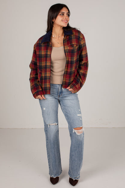 A woman modeling a red and brown plaid flannel overshirt with a dark navy collar, worn over a beige top and light-wash distressed jeans, standing against a plain light gray studio background.