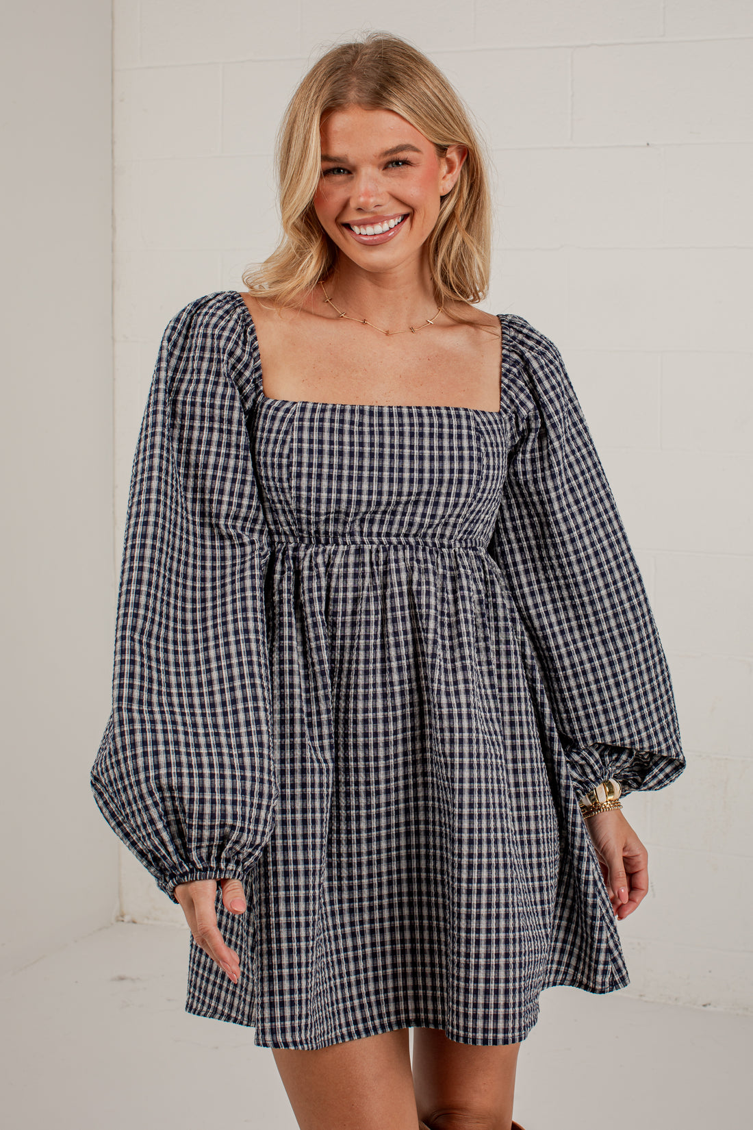A woman wearing a navy and white gingham mini dress with a square neckline, smocked bodice, and voluminous balloon sleeves, standing against a white brick wall.