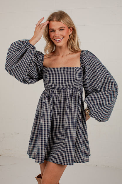 A woman modeling a navy and white gingham mini dress with voluminous puff sleeves, a square neckline, and a smocked waist, set against a white brick wall.