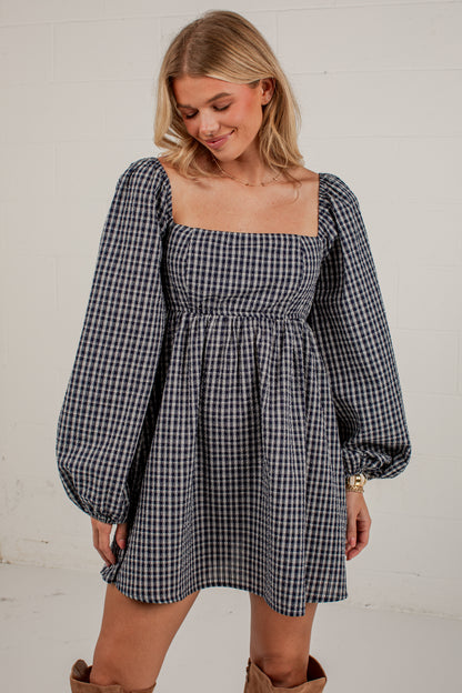 A woman wearing a navy and white gingham mini dress with puff sleeves and a square neckline, standing against a white brick wall.