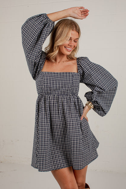 A woman modeling a navy and white gingham mini dress with voluminous puff sleeves and a square neckline, posing against a white paneled wall.
