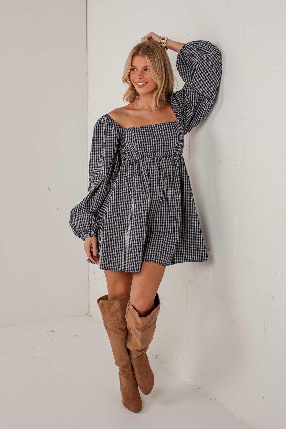 A woman wearing a black and white gingham off-the-shoulder mini dress with long puff sleeves and brown suede knee-high boots, posing against a white brick wall.