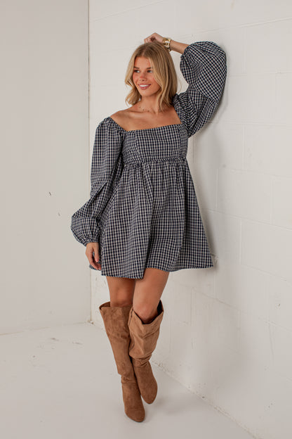A woman wearing a black and white gingham off-the-shoulder mini dress with long puff sleeves and brown suede knee-high boots, posing against a white brick wall.