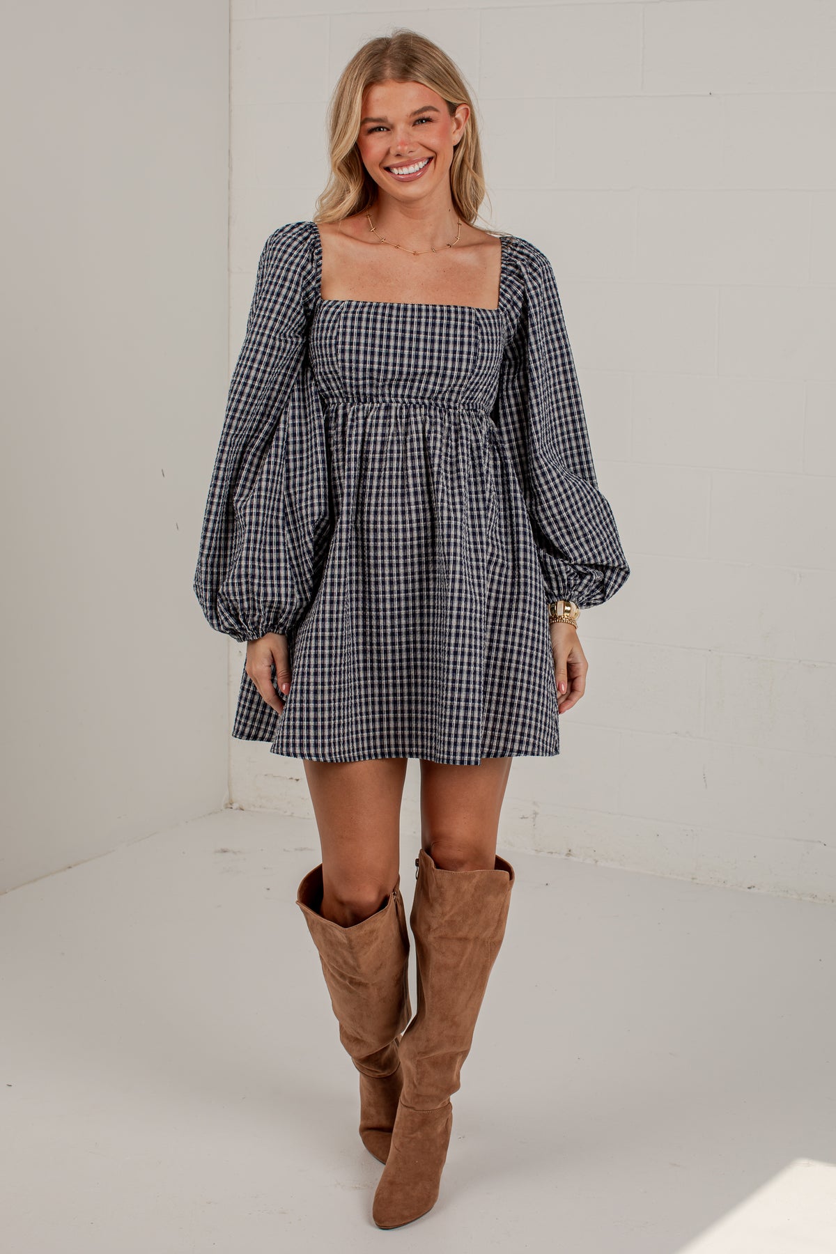 A woman modeling a black and white gingham mini dress with puff sleeves and a square neckline, paired with brown suede knee-high boots, in a bright white studio setting.