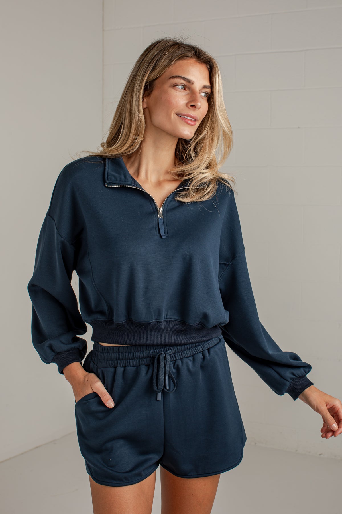 A woman wearing a matching navy blue lounge set consisting of a half-zip long-sleeve sweatshirt and drawstring shorts, standing against a light gray brick wall.