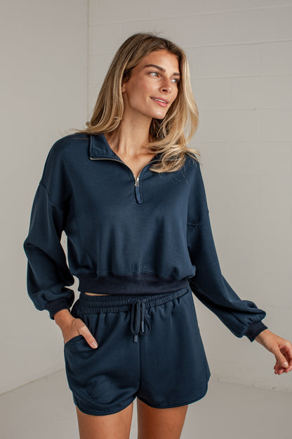 A woman wearing a matching navy blue lounge set consisting of a half-zip long-sleeve sweatshirt and drawstring shorts, standing against a light gray brick wall.