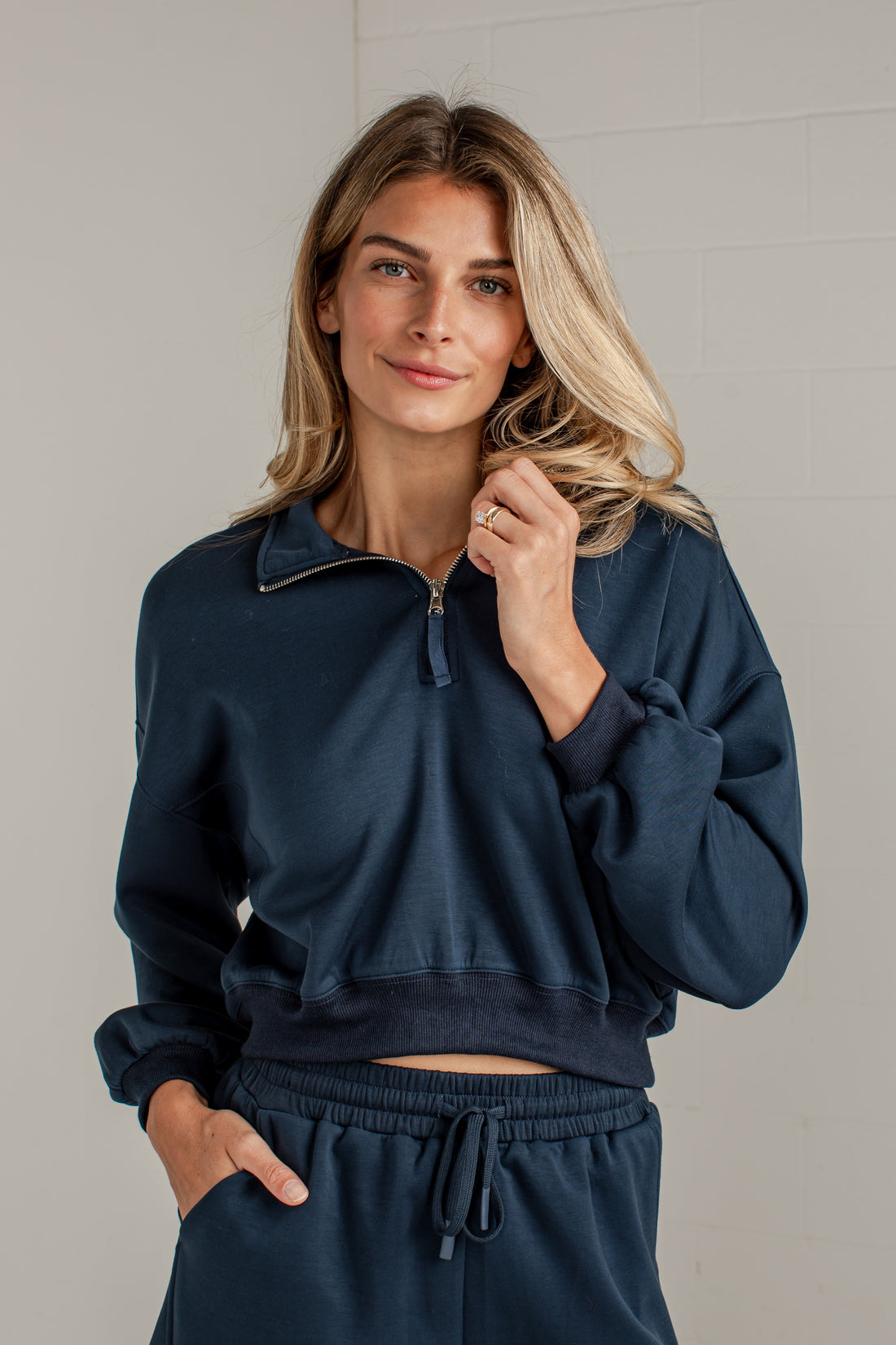 A woman wearing a navy blue cropped quarter-zip sweatshirt and matching sweatpants, standing against a light gray tiled wall.