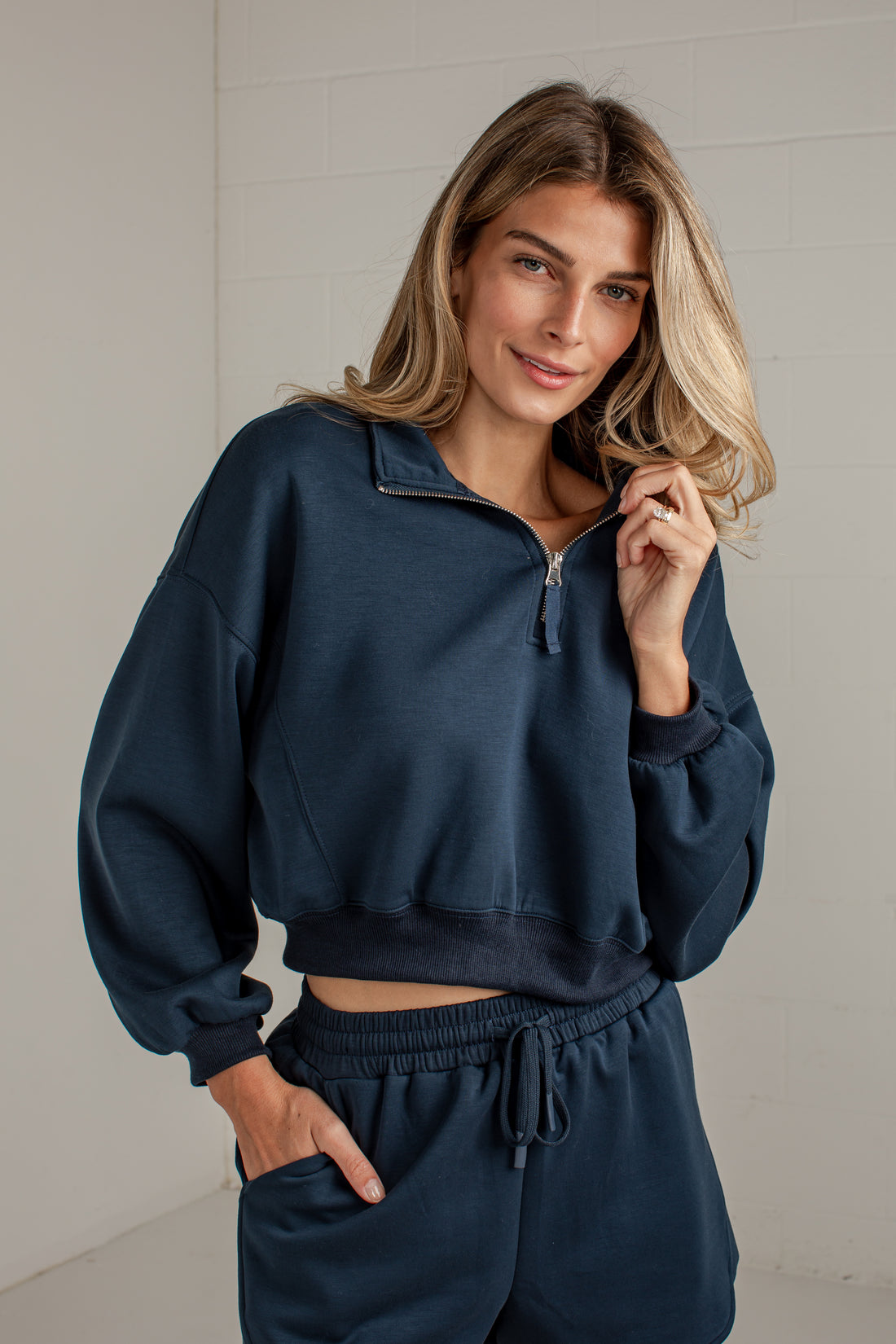 A woman wearing a navy blue cropped quarter-zip sweatshirt and matching sweatpants, standing against a light gray brick wall.