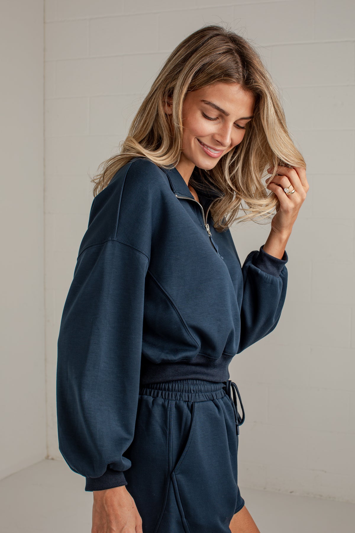 A woman wearing a navy blue long-sleeve quarter-zip sweatshirt and matching sweat shorts, standing against a light off-white paneled wall.