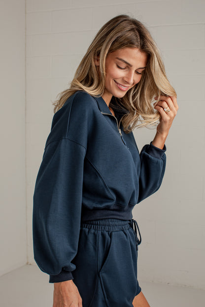 A woman wearing a navy blue long-sleeve quarter-zip sweatshirt and matching sweat shorts, standing against a light off-white paneled wall.