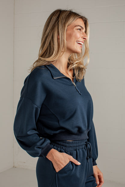 A woman wearing a navy blue long-sleeve quarter-zip sweatshirt and matching sweatpants, standing against a light gray brick wall.