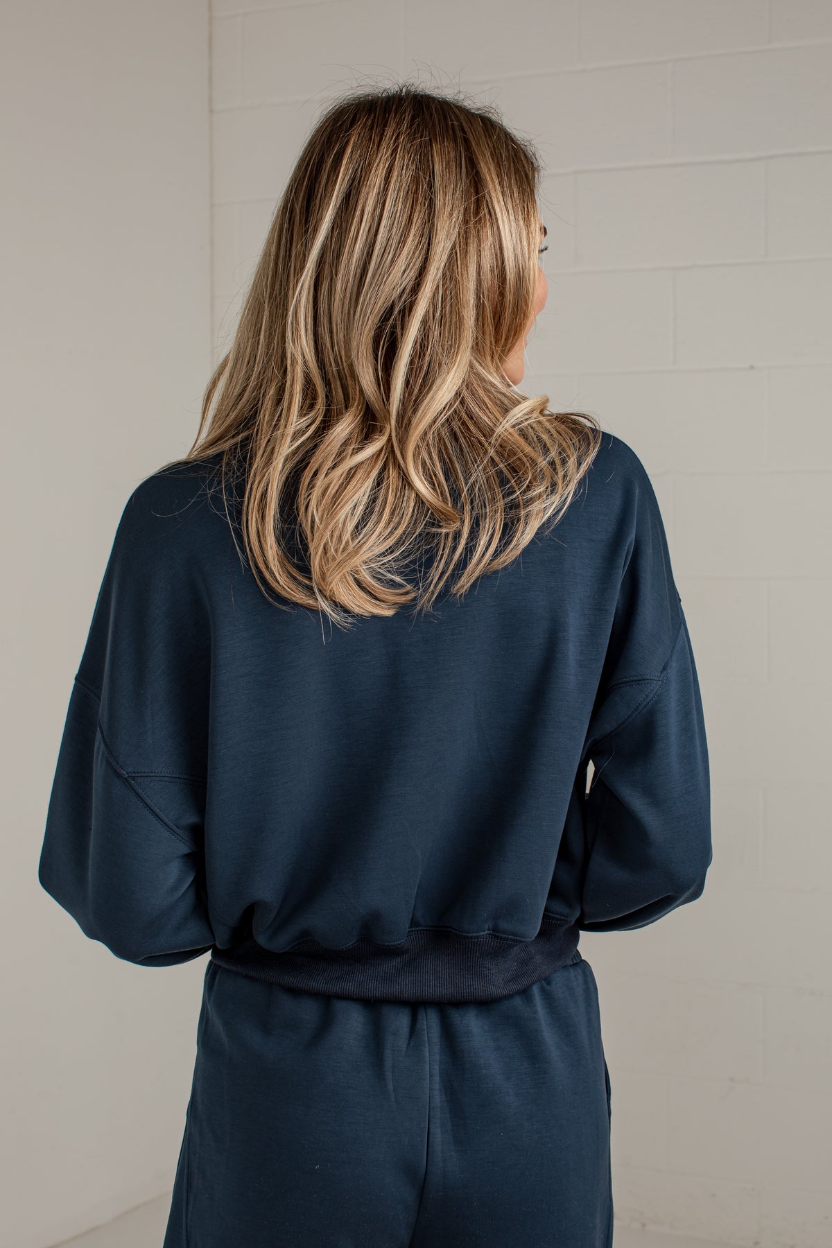 A woman viewed from behind wearing a dark navy blue sweatshirt and matching sweatpants against a light gray brick wall.