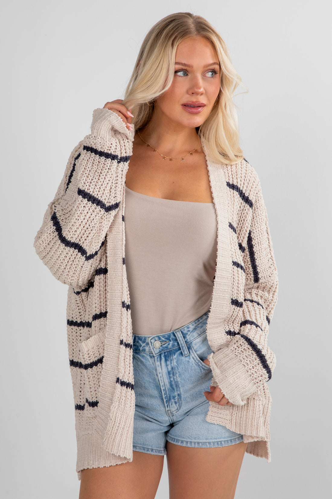 Woman wearing a beige cardigan with black stripes, white top, and denim shorts on a light gray background