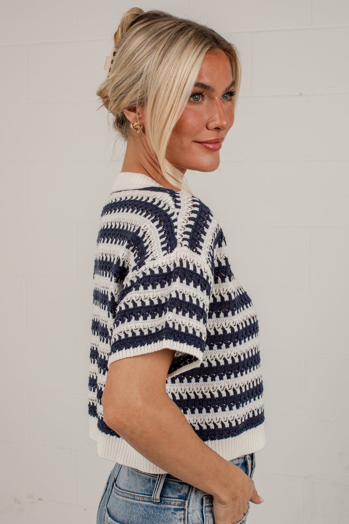A woman wearing a navy blue and off-white patterned knit short-sleeve sweater, paired with blue jeans, standing against a light gray brick wall.