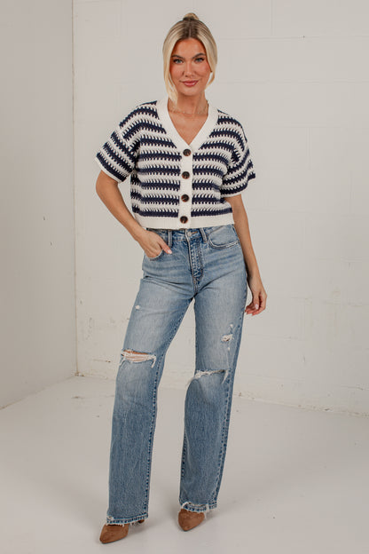 A woman modeling a short-sleeve knit cardigan with navy and white horizontal stripes, paired with light-wash distressed jeans and brown pointed-toe heels, standing against a white studio wall.