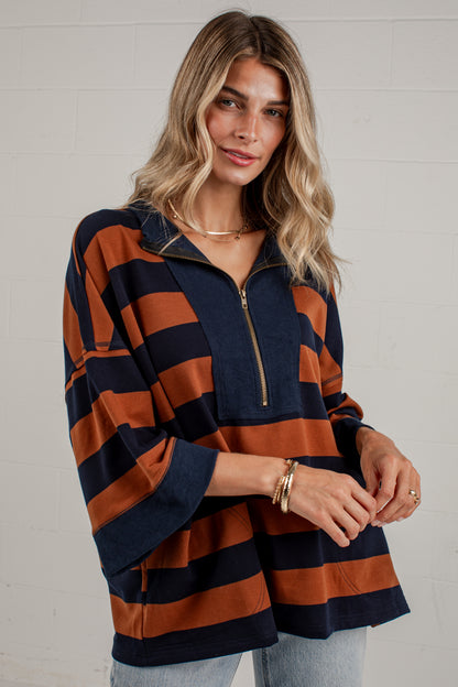 A woman wearing a navy blue and rust orange striped half-zip pullover top with wide three-quarter sleeves, paired with light-wash jeans, standing against a white brick wall.