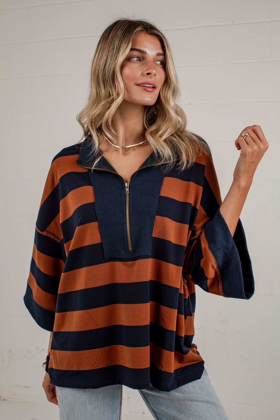 A woman wearing a loose, short-sleeve half-zip top with horizontal rust orange and navy blue stripes, paired with light-wash jeans, standing against a white brick wall.