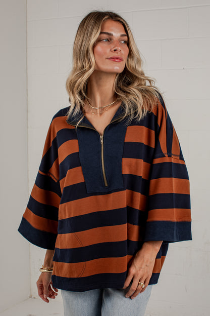 A woman wearing a loose, short-sleeve quarter-zip pullover top with horizontal navy blue and rust orange stripes, paired with light blue jeans, standing against a white brick wall.