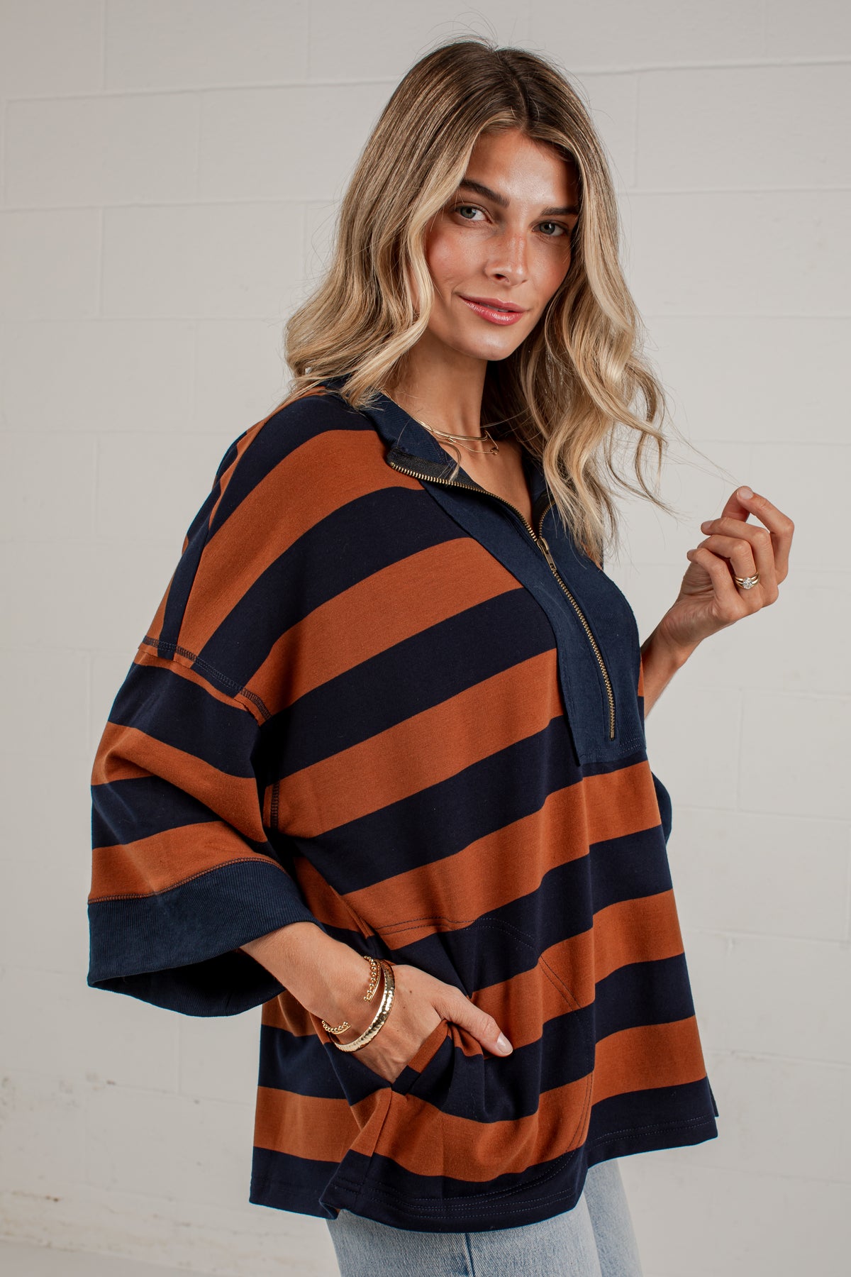 A woman wearing a loose, oversized collared top with wide horizontal stripes in rust orange and navy blue, paired with light blue jeans, standing against a white brick wall.