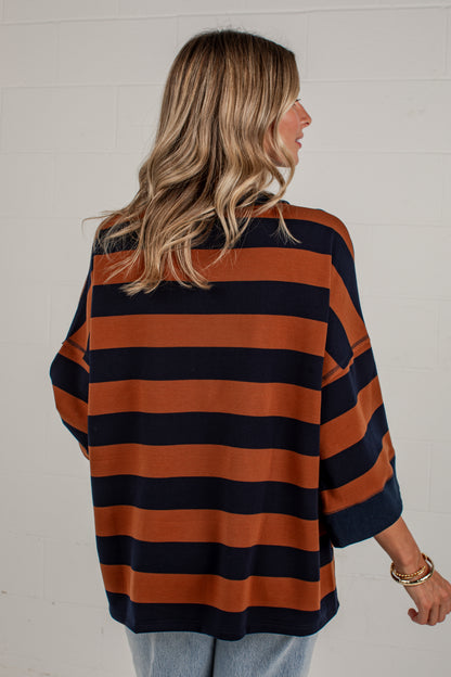 A woman shown from the back wearing an oversized long-sleeve top with horizontal rust orange and navy blue stripes, paired with light blue jeans, against a white brick wall.