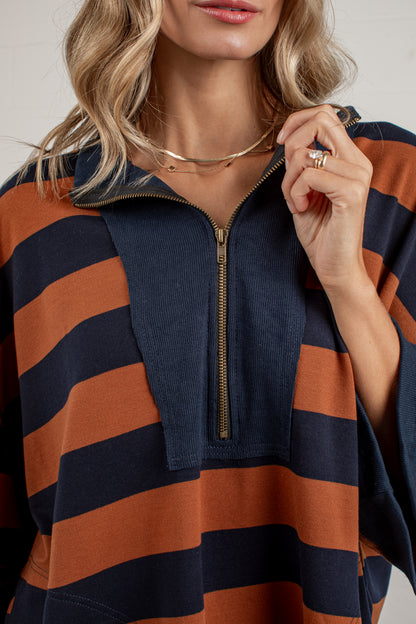 A close-up of a woman wearing a quarter-zip pullover sweatshirt with bold horizontal stripes in navy blue and rust orange. She is wearing layered gold necklaces and rings, and her blonde hair is visible against a clean, light off-white background.