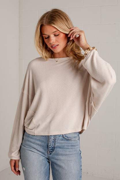 A woman wearing a light beige, oversized long-sleeve knit top with balloon sleeves and side pockets, paired with light-wash blue jeans, standing against a white brick wall.