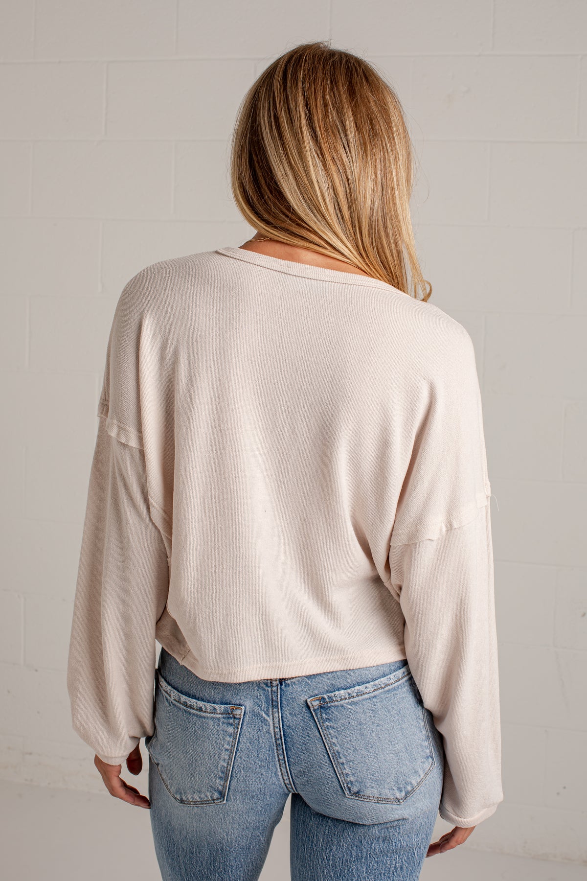 A woman viewed from behind wearing a light beige long-sleeve cropped sweater and blue jeans against a white brick wall.