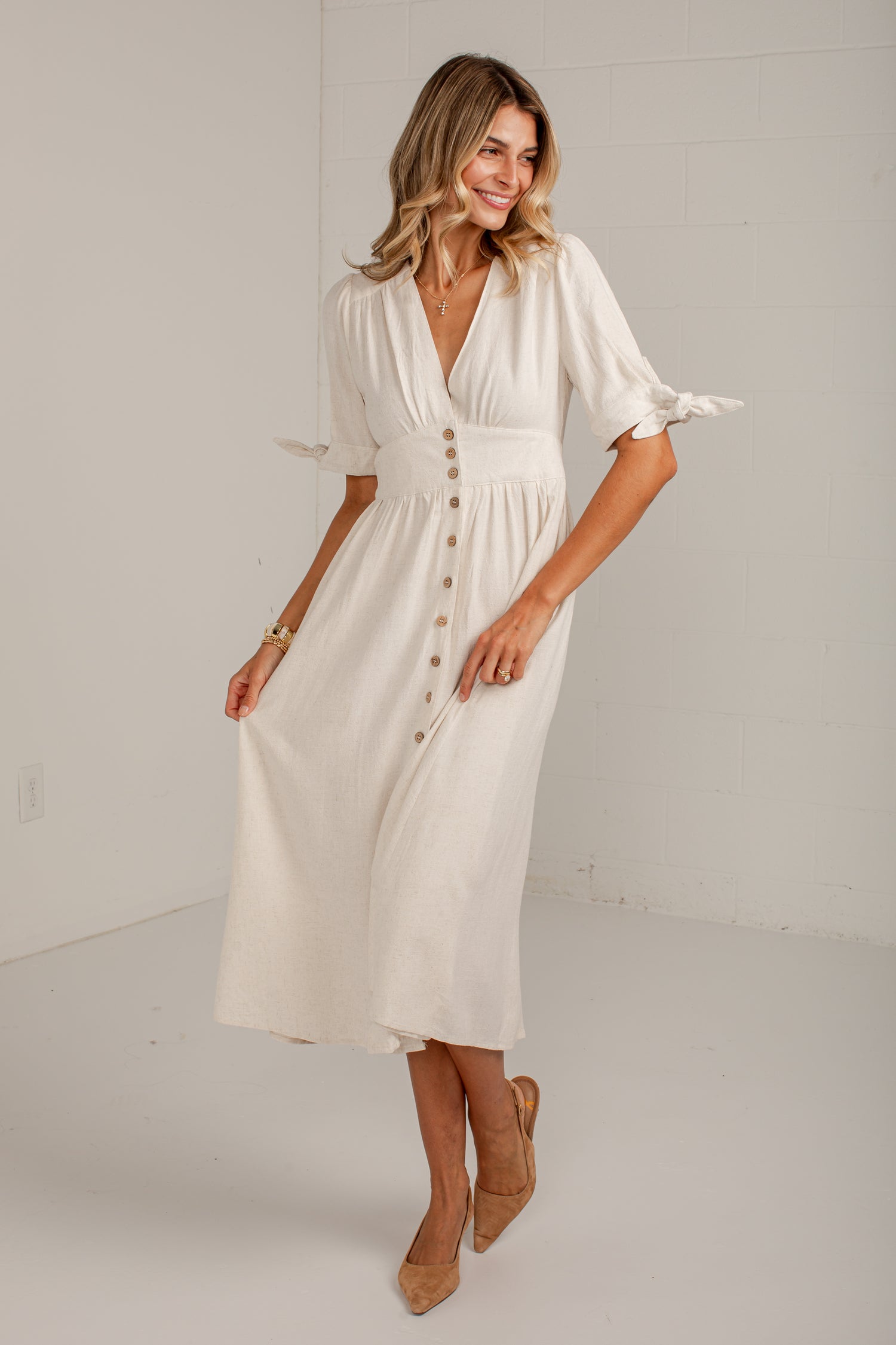 A woman modeling a cream-colored midi dress with short flutter sleeves, a V-neckline, and a button-down front, paired with tan pointed-toe heels in a bright studio setting.
