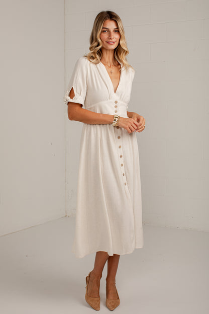 A woman modeling a cream-colored midi dress with a wrap-style V-neckline, button-down front, and cutout sleeves, standing against a plain white studio wall.