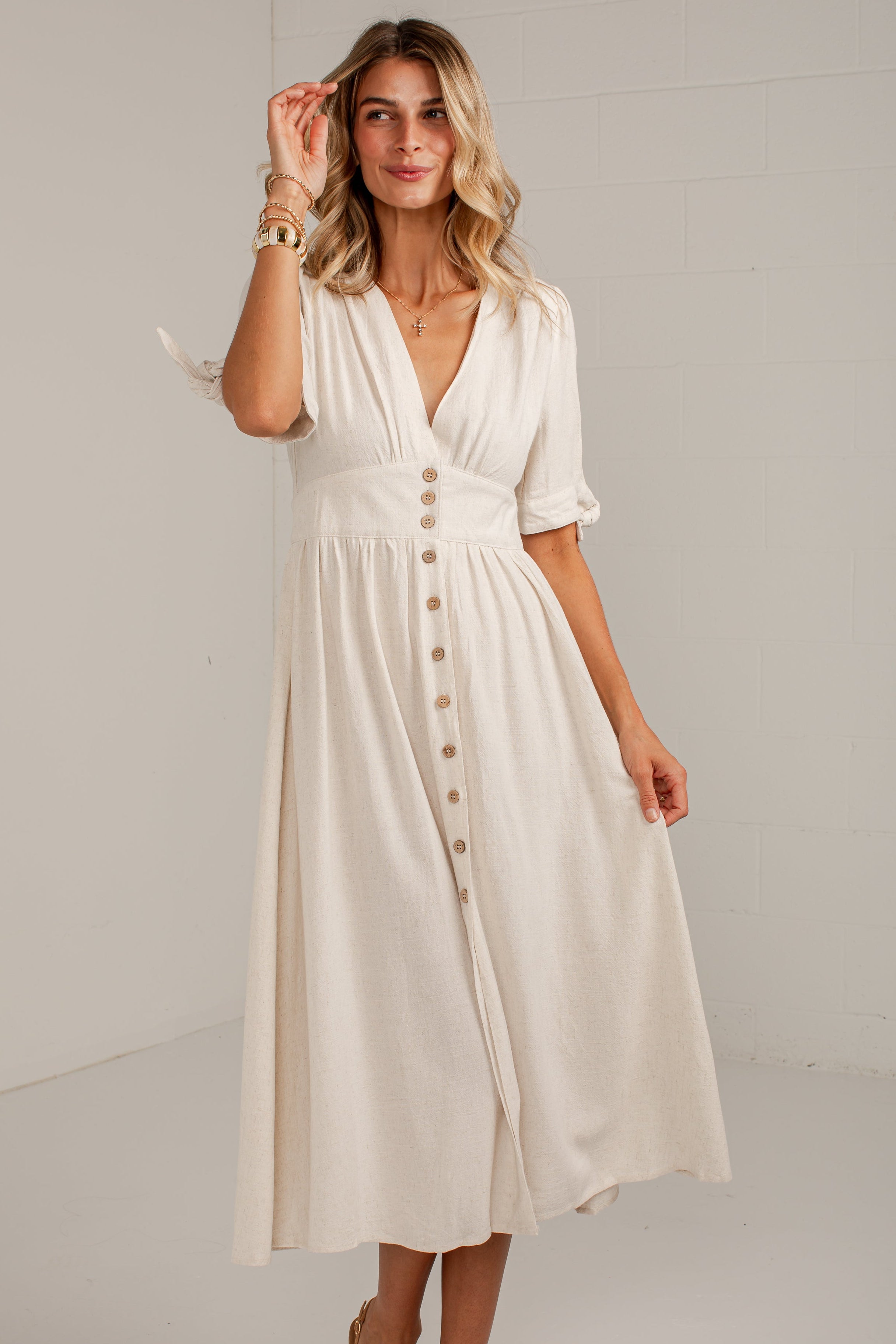 A woman modeling a cream-colored midi dress with short sleeves, a V-neckline, and a button-down front, paired with tan ankle boots in a bright studio setting.