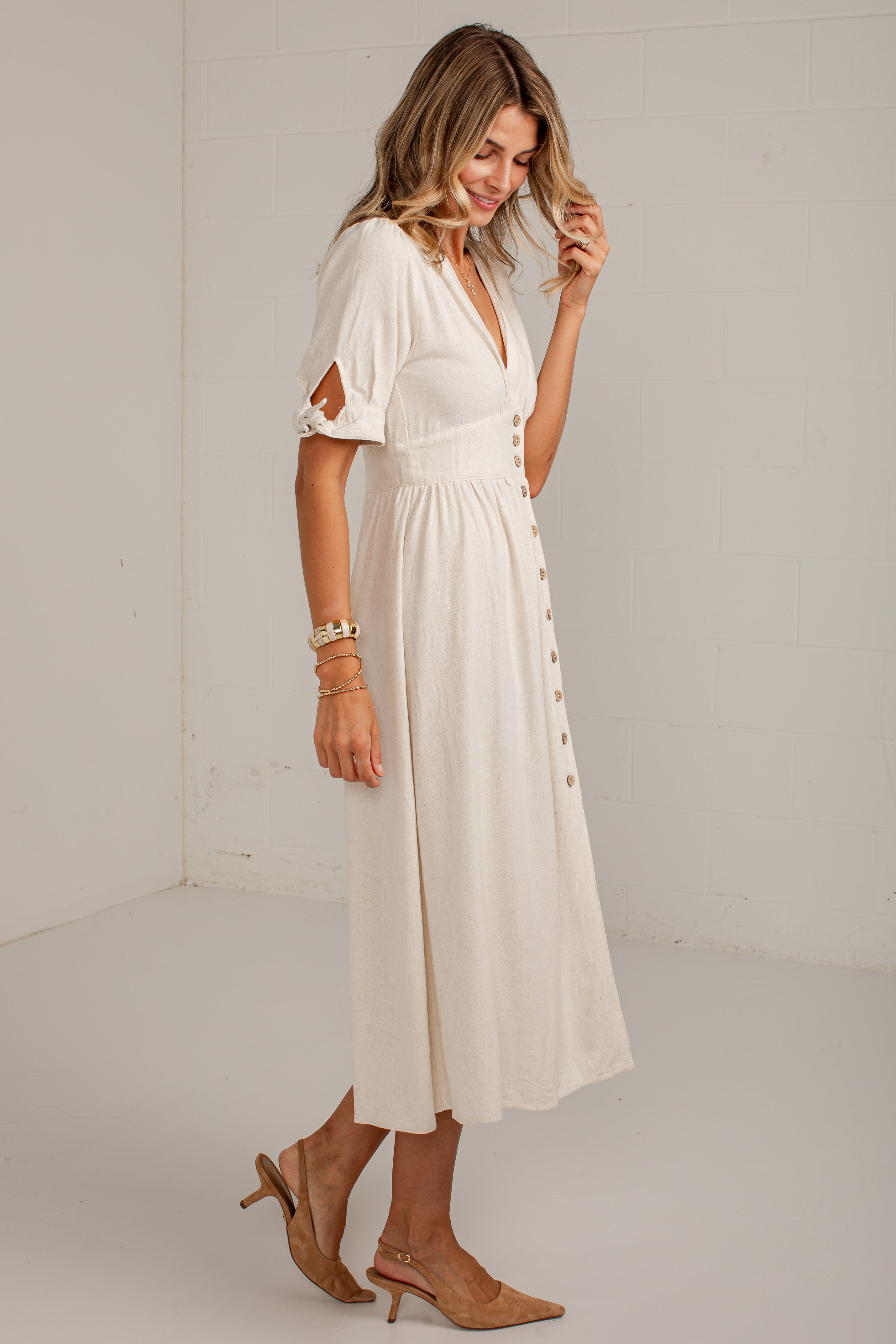 A woman modeling a cream-colored midi dress with short sleeves, a V-neckline, and side buttons, paired with tan heeled sandals in a bright studio setting.