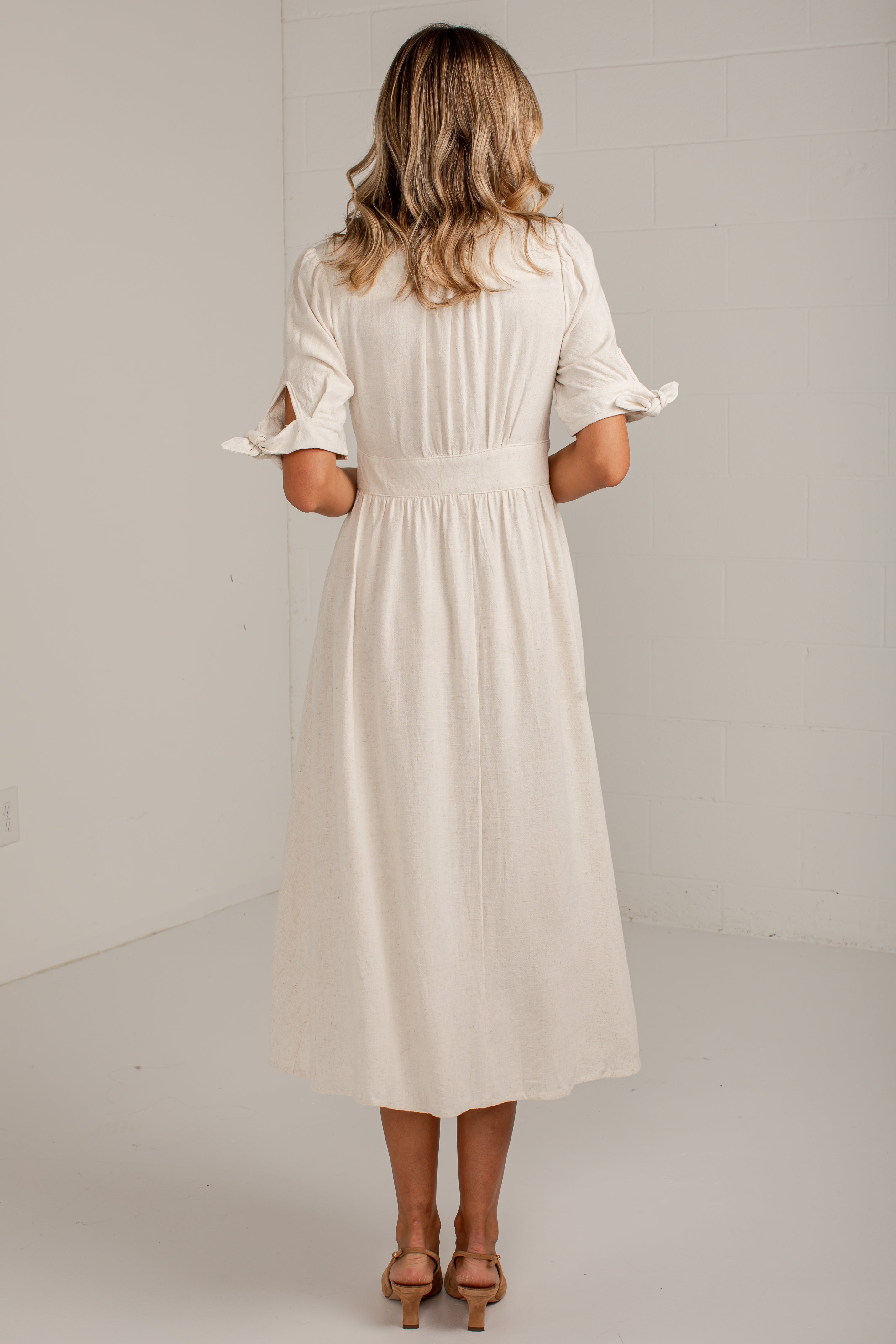 A woman viewed from behind wearing a cream-colored midi dress with ruffled sleeves and a gathered waist, paired with tan heeled sandals in a bright studio setting.