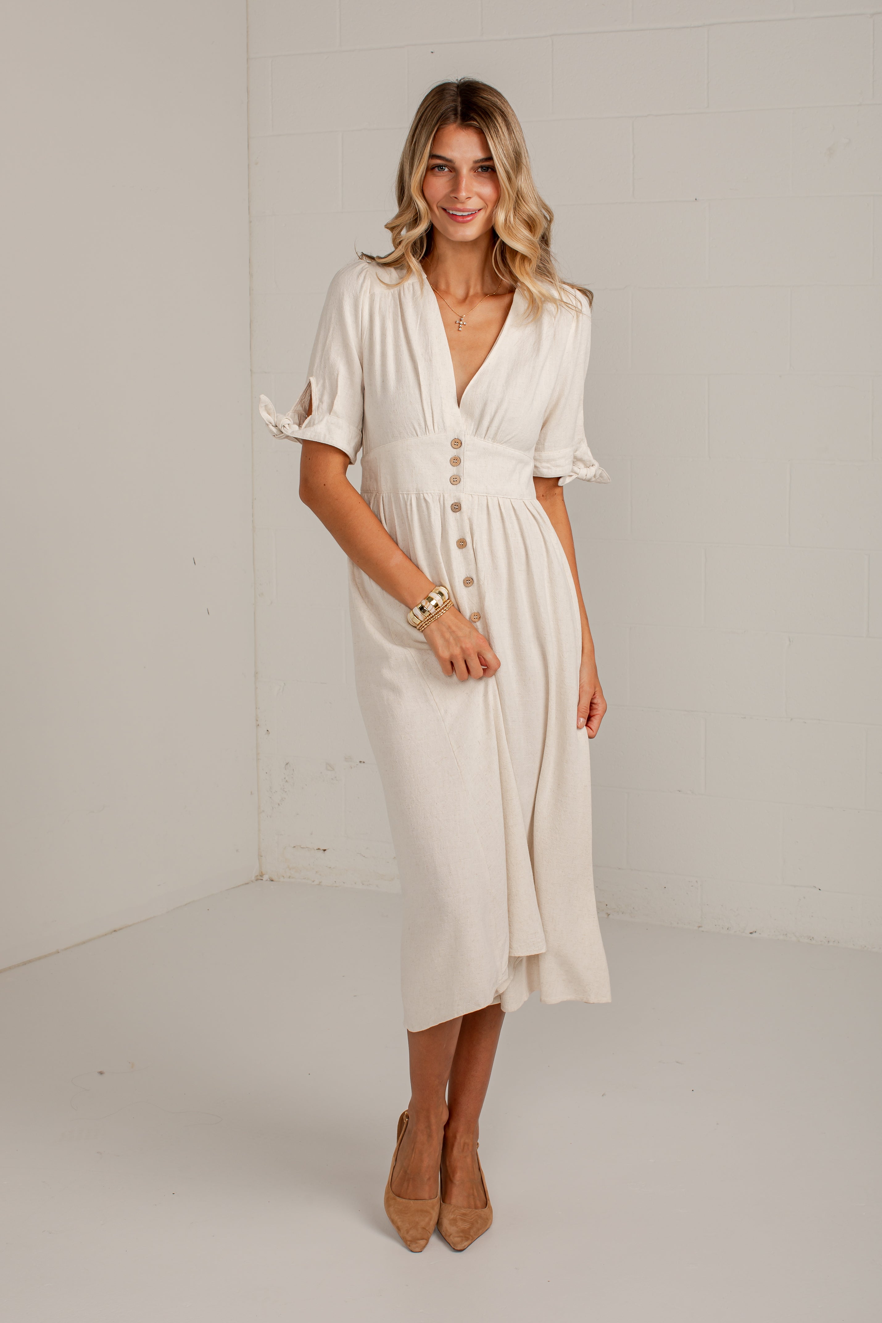 A woman modeling a cream-colored midi dress with short flutter sleeves, a V-neckline, and decorative buttons down the front, standing in a bright white studio setting.