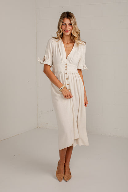 A woman modeling a cream-colored midi dress with short flutter sleeves, a V-neckline, and decorative buttons down the front, standing in a bright white studio setting.