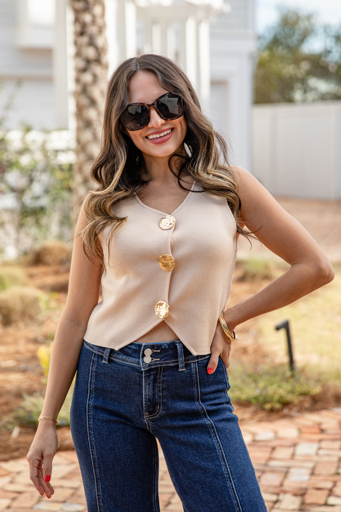 Woman wearing a beige sleeveless top with gold buttons and blue jeans outdoors.