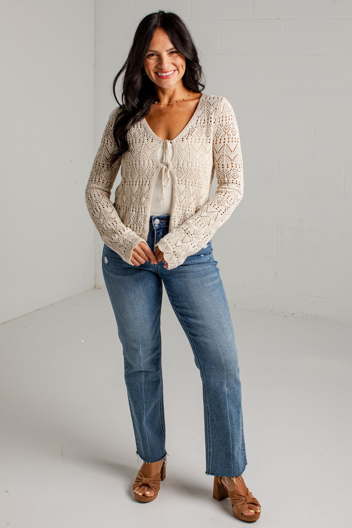 A woman modeling a cream-colored, long-sleeve crochet knit cardigan with a tie-front detail, paired with blue jeans and brown sandals, standing in a bright studio setting.