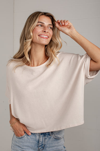 A woman wearing a light beige oversized short-sleeve t-shirt and light-wash blue jeans, posing against a light gray paneled wall.