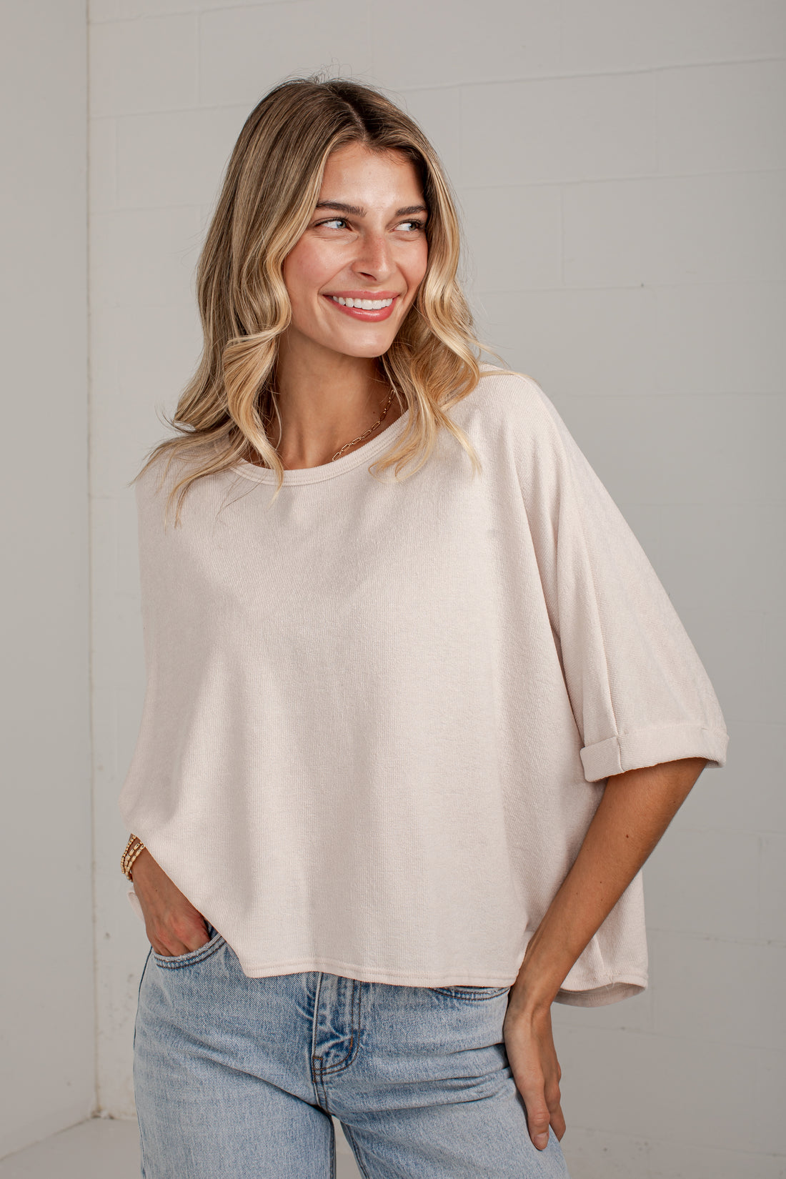A woman wearing a light beige, oversized short-sleeve top with rolled cuffs and light wash blue jeans, standing against a light gray wall.