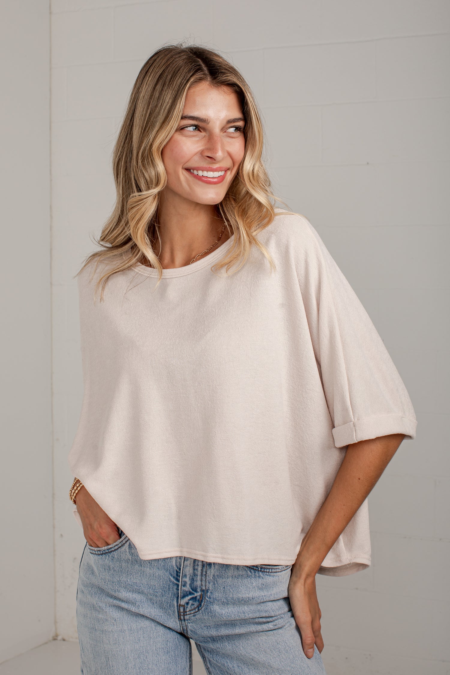A woman wearing a light beige, oversized short-sleeve top with rolled cuffs and light wash blue jeans, standing against a light gray wall.