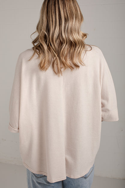 A woman viewed from behind wearing a loose, light beige knit top and blue jeans against a plain light gray wall.