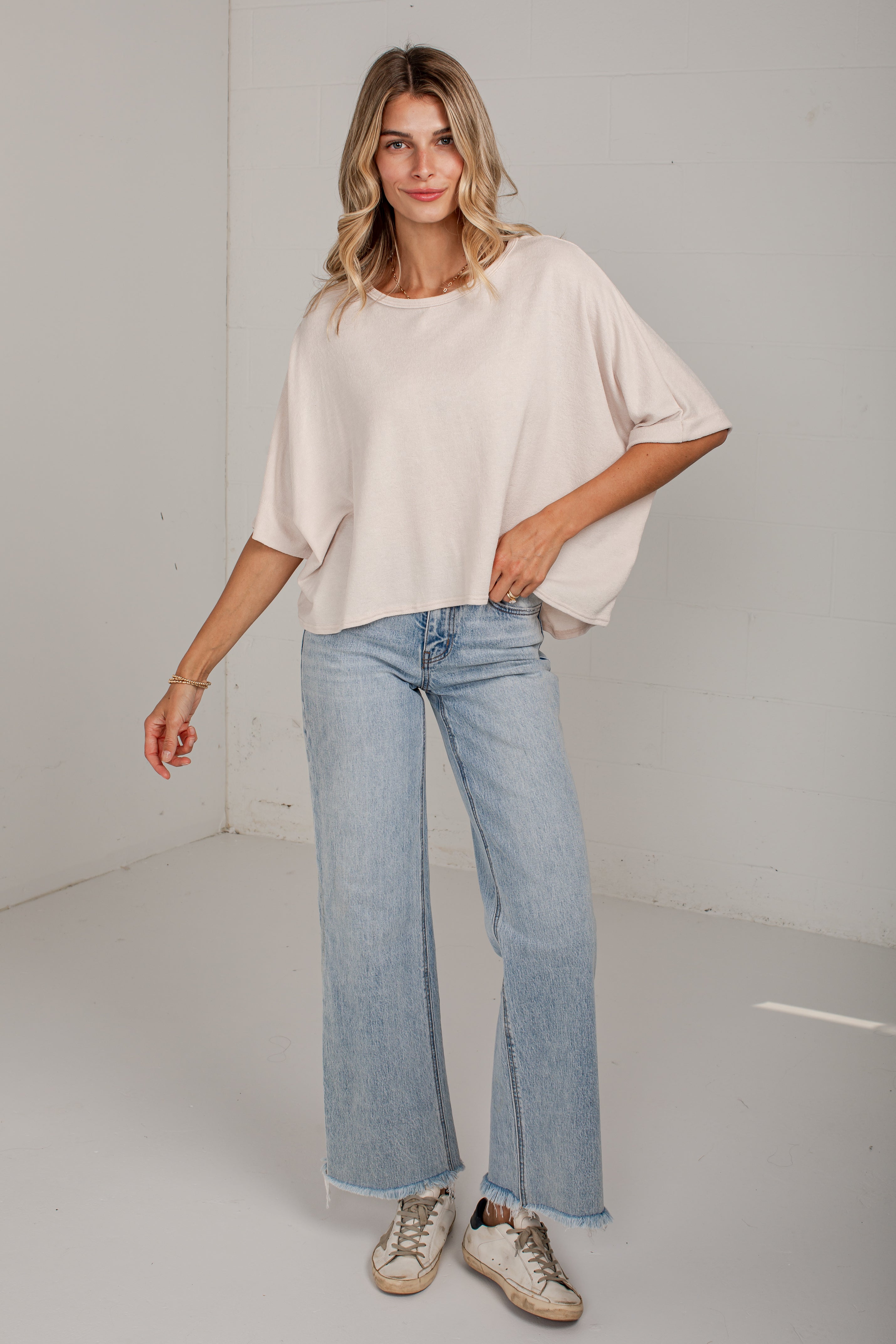 A woman modeling a light beige short-sleeve top and light blue wide-leg jeans with beige sneakers, standing in a minimalist white studio setting.