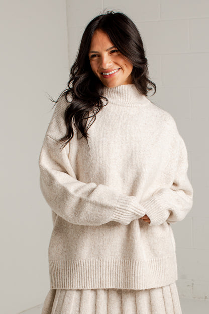 A woman wearing an oversized cream-colored knit mock neck sweater, standing against a light gray tiled wall.