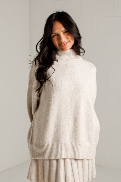A woman wearing a cream-colored knit mock neck sweater and matching pleated skirt, standing against a light gray tiled wall.