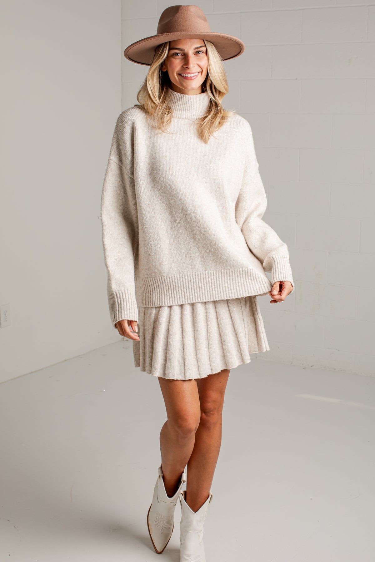 A woman modeling a cream-colored knit turtleneck sweater and matching pleated mini skirt, styled with a brown wide-brim hat and white cowboy boots against a light gray studio background.