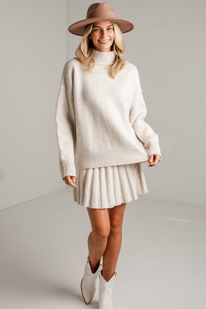 A woman modeling a cream-colored knit turtleneck sweater and matching pleated mini skirt, styled with a brown wide-brim hat and white cowboy boots against a light gray studio background.