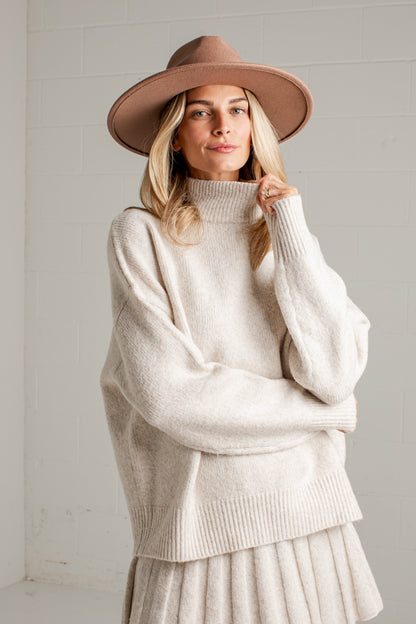 A woman wearing a cream-colored ribbed turtleneck sweater, a matching pleated skirt, and a brown wide-brim fedora hat, standing against a white brick wall.