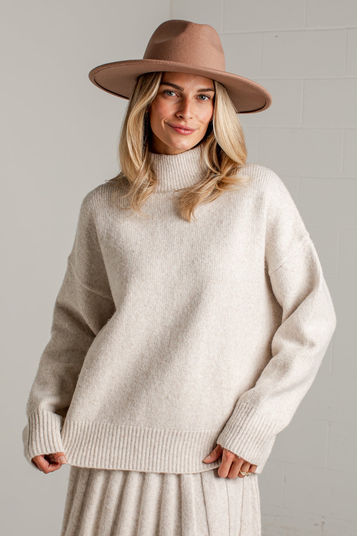 A woman wearing a cream-colored knit mock neck sweater, a matching pleated skirt, and a brown wide-brim fedora hat, standing against a light gray brick wall.